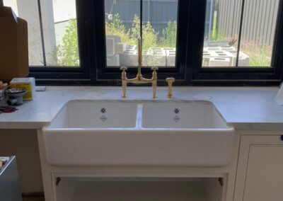 Double-basin white kitchen sink with brass faucet installed under large windows, cabinetry below. - Fosker Plumbing Western Sydney