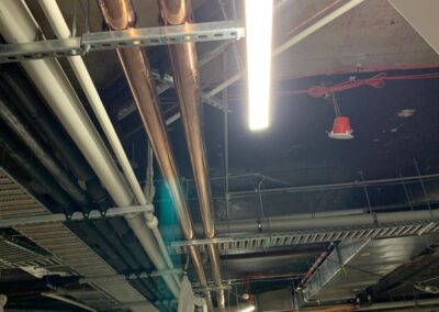 Exposed copper and PVC pipes installed on unfinished building ceiling with fluorescent light. - Fosker Plumbing Western Sydney