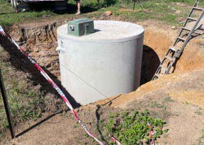 Large concrete septic tank being installed in a rural excavated site with plumbing equipment nearby.