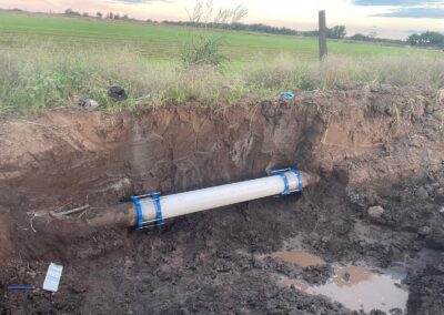 Exposed underground pipeline with blue metal clamps in muddy trench near grassy field. - Fosker Plumbing Western Sydney